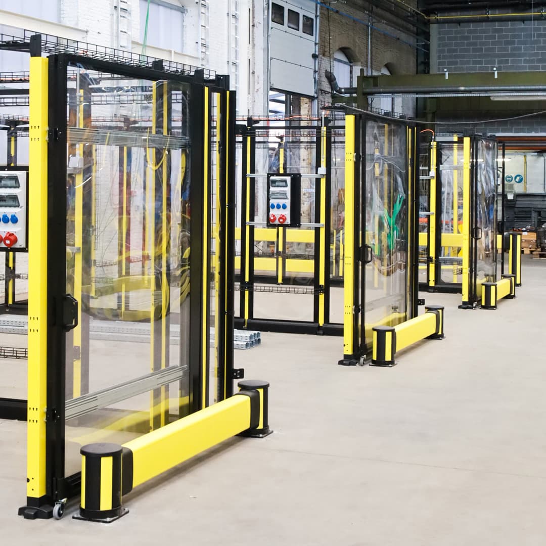 Industrial safety fencing with yellow and black metal frames and transparent panels inside a factory, enclosing machinery and control panels on a concrete floor