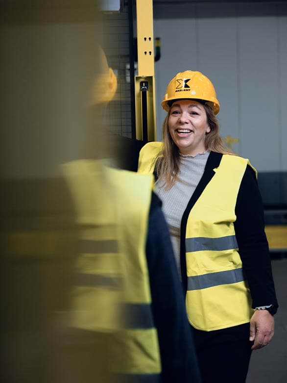 A smiling woman wearing a yellow hard hat and high-visibility vest engages in conversation with a colleague in an industrial setting. The image highlights a positive and safety-conscious work environment, reinforcing Axelent’s commitment to workplace safety and teamwork.