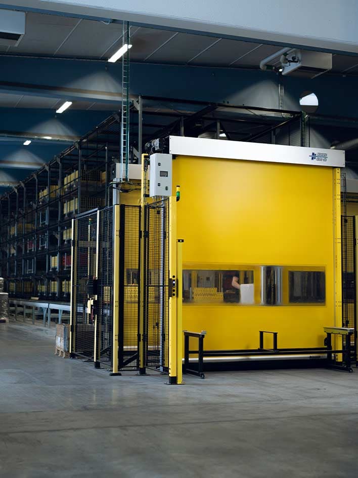 An industrial safety barrier with a yellow automated roll-up door and protective fencing, securing access to a warehouse or production area. The image highlights the importance of selecting machine guards that balance safety, efficiency, and accessibility in industrial environments.