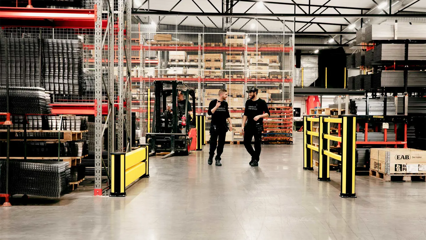 A well-organized industrial warehouse with employees walking and discussing, highlighting a good and healthy work environment. Safety barriers and structured storage emphasize workplace safety and efficiency.